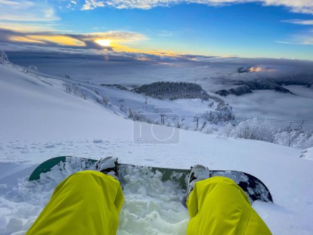 Téléchargez les photos : POV: Looking at the golden sunset while sitting in the fresh powder snow during a snowboarding trip in the Julian Alps. Stopping during an offpiste snowboarding session to watch the scenic sunrise. - en image libre de droit