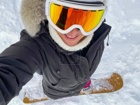 Foto de VERTICAL, SELFIE: Young Caucasian woman smiles while snowboarding off piste in the pristine Julian Alps. Cheerful female snowboarder takes a selfie while riding in backcountry covered in fresh snow - Imagen libre de derechos