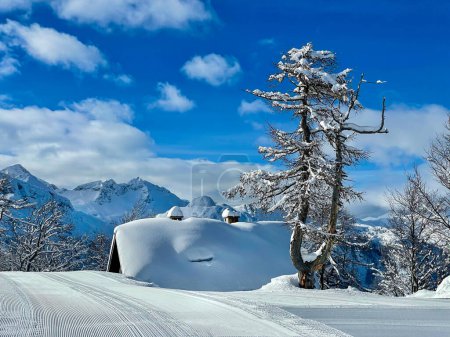 Foto de Empty groomed ski resort slope runs past a large wooden cottage in the Julian Alps covered in fresh snow. Scenic shot of the stunning alpine landscape in the Slovenian mountains on a sunny winter day. - Imagen libre de derechos