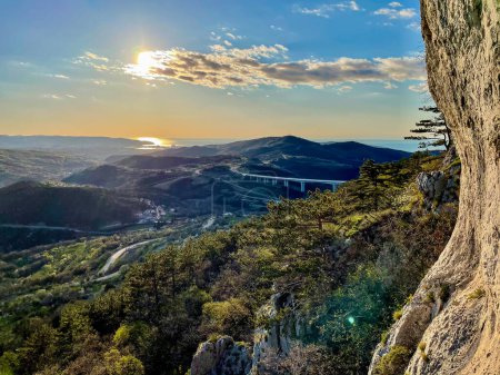Téléchargez les photos : LENS FLARE: Spectacular view of the Crni Kal landscape on a sunny summer day. Rocky climbing wall in rural Slovenia overlooks the lush landscape near the seaside and a highway viaduct crossing it. - en image libre de droit