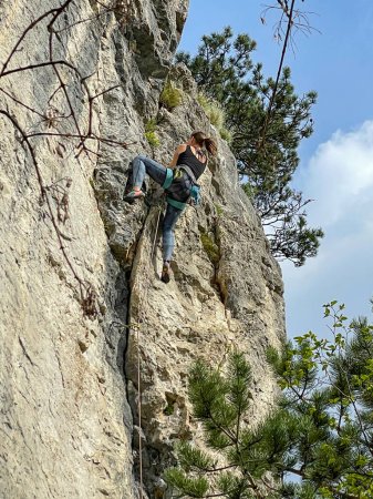 Photo for VERTICAL, BOTTOM UP: Young woman top rope rock climbs up a towering cliff in Slovenia. Athletic female tourist is climbing up a rocky wall in Crni Kal. Extreme rock climber in action on a sunny day. - Royalty Free Image