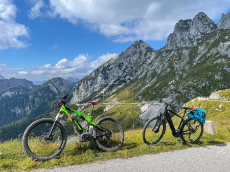 Téléchargez les photos : Two electric bicycles are parked next to an empty mountain road in Slovenia on a sunny summer day. Scenic shot of two e-bikes parked at the side of the road overlooking the gorgeous Julian Alps. - en image libre de droit