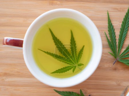 Photo for Fragrant indica marijuana leaf steeps in boiling hot water inside a brown mug sitting on the wooden countertop. Sativa weed leaves are being used to brew delicious medicinal tea. - Royalty Free Image