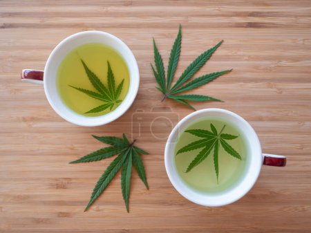 Téléchargez les photos : Fragrant indica marijuana leaf steep in hot water inside of two brown mugs sitting on the wooden countertop. Sativa weed leaves are being used to brew delicious medicinal tea. - en image libre de droit