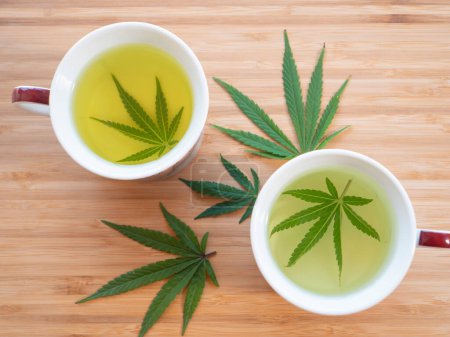Photo for Homegrown indica weed leaves are being used to prepare therapeutic tea. Detailed shot of two teacups filled with illegal homebrewed marijuana tea. Preparing tea on the wooden table. - Royalty Free Image