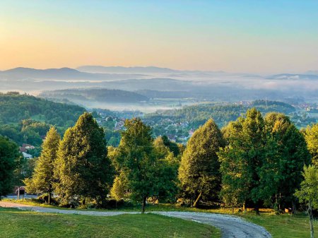 Téléchargez les photos : Mist covering the idyllic valley in Slovenian countryside clears up at golden sunrise. Scenic shot of the lush green countryside and the village of Sticna on a sunny spring morning. Idyllic landscape. - en image libre de droit