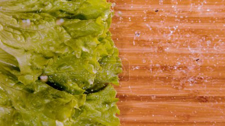 Téléchargez les photos : TOP DOWN, CLOSE UP, COPY SPACE: Wet romaine lettuce leaves fall onto the empty wooden chopping board. Cinematic shot of glassy water droplets flying as wet iceberg hits the chef's cutting board. - en image libre de droit