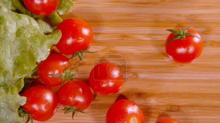 Foto de MACRO, DOF: Cinematic shot of shiny wet tomatoes falling and rolling around a wooden countertop. Cleaned salad ingredients land on the kitchen counter. Organic lettuce and tomatoes on cutting board. - Imagen libre de derechos