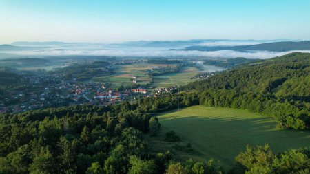 Téléchargez les photos : AERIAL: Mist covering the idyllic valley in Slovenian countryside clears up at spring sunrise. Scenic drone shot of the lush green countryside and the village of Sticna on a cold spring morning. - en image libre de droit