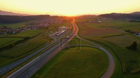 Foto de AERIAL, LENS FLARE: Volando a lo largo de una carretera concurrida y hacia la hermosa puesta de sol mientras los coches y camiones conducen por la carretera escénica. Los rayos de sol dorados de la noche de verano brillan en la autopista de Eslovenia. - Imagen libre de derechos