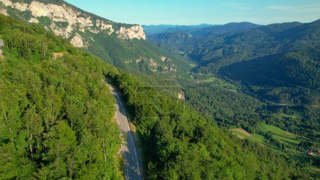 Téléchargez les photos : AÉRIEN : Une route de montagne pittoresque traverse la forêt verdoyante qui recouvre le paysage pittoresque de la Slovénie rurale. Vue aérienne d'une route vide dans des bois verdoyants élevés dans les montagnes de Slovénie. - en image libre de droit
