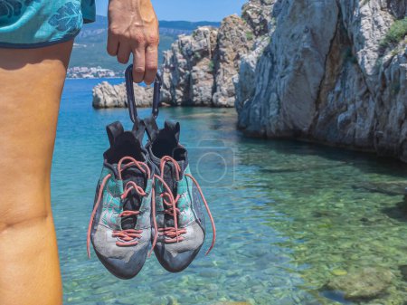 Foto de CLOSE UP: Held climbing shoes with climbing boulder above seawater in background. Young woman holding sports shoes tied with a carabiner before going deep water solo climbing at the beautiful seaside. - Imagen libre de derechos