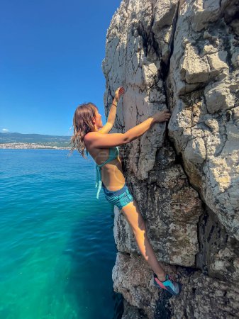Foto de CLOSE UP: Sporty lady in swimsuit rock climbing on a boulder above blue seawater. Picturesque seaside location for sports activity. Young woman deep water solo climbing at rocky seashore in summer. - Imagen libre de derechos