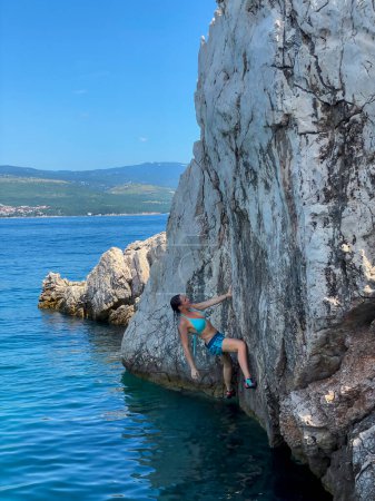 Téléchargez les photos : Young woman checking options for climbing a rock boulder above blue seawater. Amazing seaside climbing spot with female climber at deep water solo climbing. Picturesque location for sports activity. - en image libre de droit