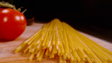 Foto de Sheaf of uncooked spaghetti pasta placed on wooden countertop. Italian pasta ready for cooking and preparing delicious meal. Pasta and tomato on wooden surface with black background. - Imagen libre de derechos
