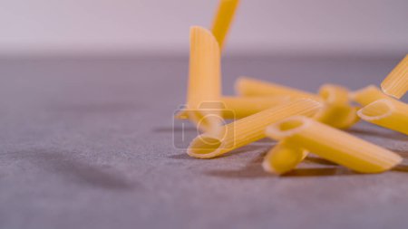 Téléchargez les photos : Scattered pasta pennette rigate in mid-air on grey background. Right side throw of falling penne pasta in mid-air. Detailed view of flying Italian uncooked pasta rigatoni. - en image libre de droit