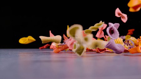 Téléchargez les photos : Pasta in variety of shapes and colours falling on gray surface. Close up of scattered coloured pasta mixture caught in motion. Detailed view of several sorts of Italian raw pasta. - en image libre de droit