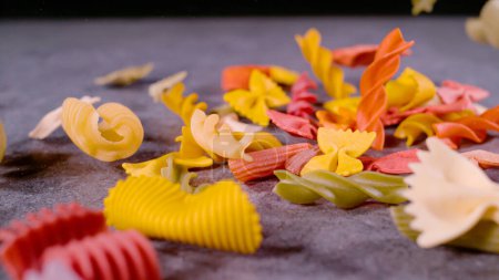 Téléchargez les photos : Pasta of different shapes and colours dropped on grey surface. Close up of scattered coloured pasta mixture with shallow focus. Detailed view of several types of Italian raw pasta. - en image libre de droit