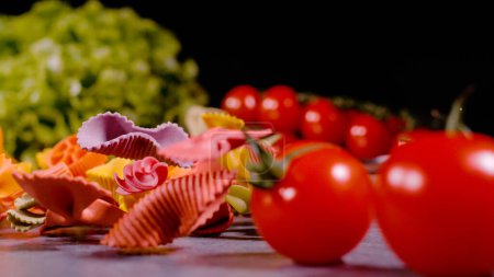 Photo for Pasta in variety of shapes and colours between ripe red tomatoes in foreground and green lettuce in background. Several types of Italian raw pasta among vegetables on grey countertop. - Royalty Free Image