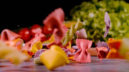 Téléchargez les photos : Pasta in variety of shapes and colours falling on grey kitchen countertop with ripe red tomatoes and green lettuce in background. Dropped Italian colored raw pasta in shallow focus. - en image libre de droit