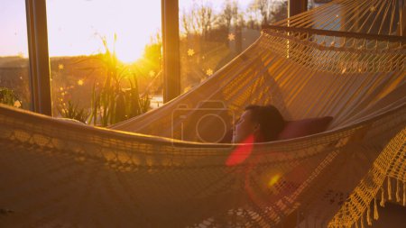 Foto de CLOSE UP: Young woman resting peacefully in a cozy hammock while sun is setting. Self care treatment to regain health. Late afternoon rest during sick leave in home living room while sun is setting. - Imagen libre de derechos
