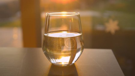 Foto de CLOSE UP: Fresh water in glass jar backlit with golden sunlight placed on table. Sun reflecting from drinking glass filled with water. Transparent and colourless fluid in a glass jar. - Imagen libre de derechos
