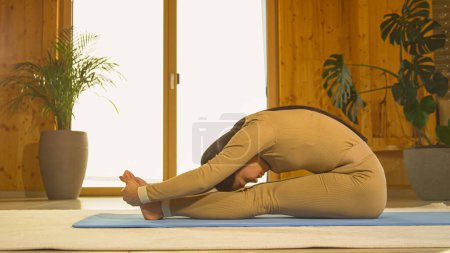 Téléchargez les photos : Athletic young Asian woman practicing seated forward fold yoga posture. Young female person doing body stretching on blue yoga mat. Healthy indoor activity for relaxation and flexibility. - en image libre de droit