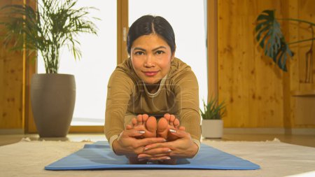 Photo for Young Filipino woman stretching her legs and working out at home. Asian female person doing seated forward fold yoga pose while looking at camera. Healthy leisure activity for vitality. - Royalty Free Image