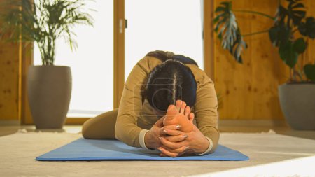 Photo for Front view of Asian woman practicing seated head to knee yoga posture. Young female person doing body stretching on blue yoga mat. Healthy indoor activity for relaxation and flexibility. - Royalty Free Image