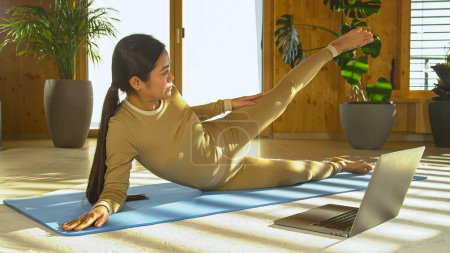 Photo for Sporty Asian woman attending online pilates workout class using laptop. Beautiful Philippine woman in sportswear smiling while exercising pilates to gain strength in thigh and hip muscles. - Royalty Free Image