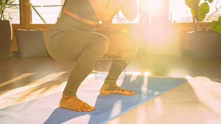 Foto de Sporty young woman performing squats at home workout in golden light. Fit lady in sportswear during bodyweight training doing exercises for leg and lower back muscles and body stability. - Imagen libre de derechos