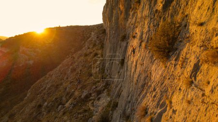 Foto de AERIAL: Escaladora subiendo por la pared con destello de sol dorado en el fondo. Mujer escalando en medio de la pared durante el atardecer. Actividad al aire libre adrenalina en un hermoso entorno natural. - Imagen libre de derechos
