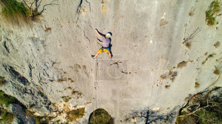 Photo for AERIAL: Young male rock climber lead climbing and using quickdraw to attach rope. Male climber in the middle of sunlit limestone wall hooking the rope. Outdoor sport activity in natural environment. - Royalty Free Image