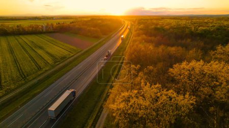 Téléchargez les photos : AÉRIAL : Convoi de camions cargo sur une autoroute rurale dans une belle lumière dorée. Faible circulation sur une autoroute qui traverse un beau paysage sous une lumière magnifique. Transport routier pour la livraison de marchandises. - en image libre de droit