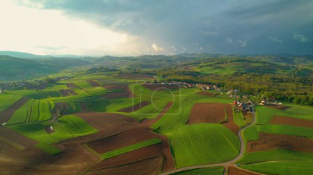 Foto de AERIAL: Picturesque hilly area with meadows, fields and villages in morning light. Charming countryside with lovely pattern of pastures and farm fields. Idyllic landscape bathing in afternoon light. - Imagen libre de derechos