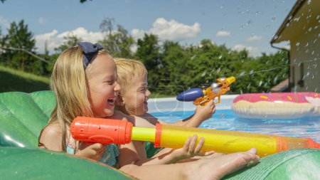 Téléchargez les photos : FERMER : Frère et sœur souriants profitant du jeu de billard avec des blasters. Enfants heureux riant tout en flottant dans la piscine. Eau pour se rafraîchir pendant les journées chaudes et ensoleillées de l'été. - en image libre de droit