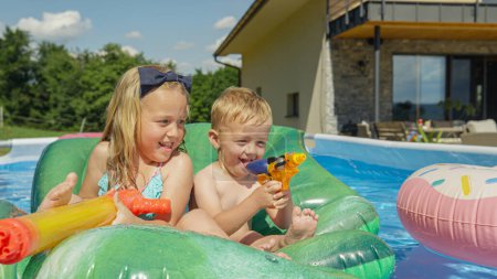 Téléchargez les photos : FERMER : Deux enfants heureux souriant et s'amusant dans la piscine de l'arrière-cour. Soeur et frère profitant de la fête de piscine flottant sur des jouets gonflables par une journée d'été ensoleillée. Jeux d'eau pour les jours chauds - en image libre de droit