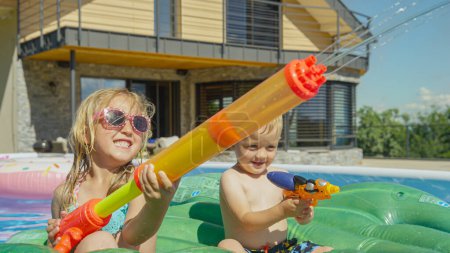 Foto de CLOSE UP: Happy brother and sister having fun at water fight in the garden pool. Water games for hot summer days. Children enjoying and having fun while playing in the swimming pool at home backyard. - Imagen libre de derechos