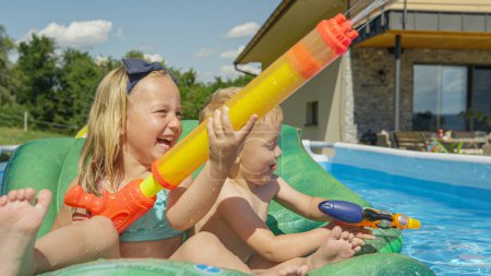 Foto de CLOSE UP: Happy and smiling children enjoying at water fight in the garden pool. Water games for hot summer days. Children enjoying and having fun while playing in the swimming pool at home backyard. - Imagen libre de derechos