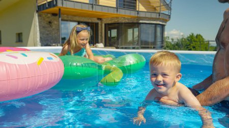 Foto de CLOSE UP: Smiling little boy floating in backyard swimming pool learning to swim. Cheerful toddler at his first swimming swings in home pool. Refreshing water family activities on hot summer days. - Imagen libre de derechos