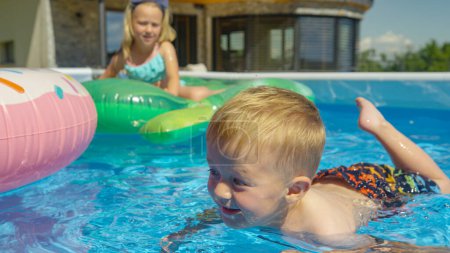 Foto de CLOSE UP: Cute little boy floating in backyard swimming pool and learning to swim. Cheerful toddler at his first swimming swings in home pool. Refreshing water family activities on hot summer days. - Imagen libre de derechos