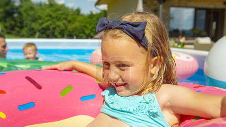 Foto de CLOSE UP: Smiling little girl enjoying floating on inflatable donut in the pool. Cheerful and smiling kid having fun during summer activities and refreshment in home garden swimming pool on a hot day. - Imagen libre de derechos