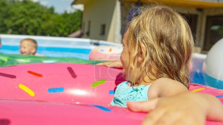 Foto de CERRAR: Adorable chica disfrutando flotando en la rosquilla inflable en la piscina. Alegre y sonriente niño divirtiéndose durante las actividades de verano y refresco en casa jardín piscina en un día caluroso. - Imagen libre de derechos