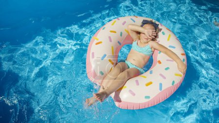 Téléchargez les photos : TOP DOWN: Little girl in pool splashing water on a floatie donut hiding her face. Cute girl covering her face with hand while floating and playing in backyard pool. Fun activity for summer holidays. - en image libre de droit