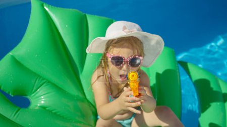 Foto de TOP DOWN Little girl sitting on a floatie with water gun pointing towards camera. Cheerful little girl floating in backyard swimming pool and having fun with water blaster spraying in camera direction - Imagen libre de derechos