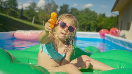 Foto de CLOSE UP: Adorable girl sitting on a floatie in garden pool holding a water gun. Playful little girl floating in backyard swimming pool and having fun with water blaster spraying directly into camera - Imagen libre de derechos