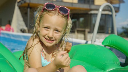 Photo for PORTRAIT: Sweet girl with ice cream bar floating in the garden pool on a hot day. Adorable girl eating chocolate ice cream in backyard swimming pool. Complete refreshment on a warm sunny summer day. - Royalty Free Image