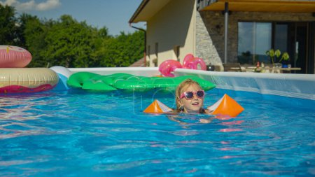 Téléchargez les photos : FERMER : Petite fille mignonne avec des lunettes de soleil nageant dans la piscine avec des flotteurs à bras. Adorable fille pratiquant la natation avec des ailes d'eau dans la piscine de jardin. Savourez un rafraîchissement lors d'une journée d'été chaude et ensoleillée. - en image libre de droit