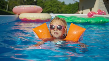 Foto de PORTRAIT: Cheerful girl with sunglasses swimming in the pool with water wings. Cute girl practicing swimming with arm floats in garden pool. Enjoyful and fun refreshment on a hot and sunny summer day. - Imagen libre de derechos