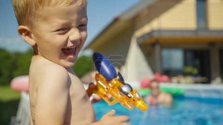 Foto de CLOSE UP Cheerful little boy having fun at water fight near the swimming pool. Playful holiday moment of a child enjoying at water game. Fun and refreshing outdoor activities for hot sunny summer days - Imagen libre de derechos
